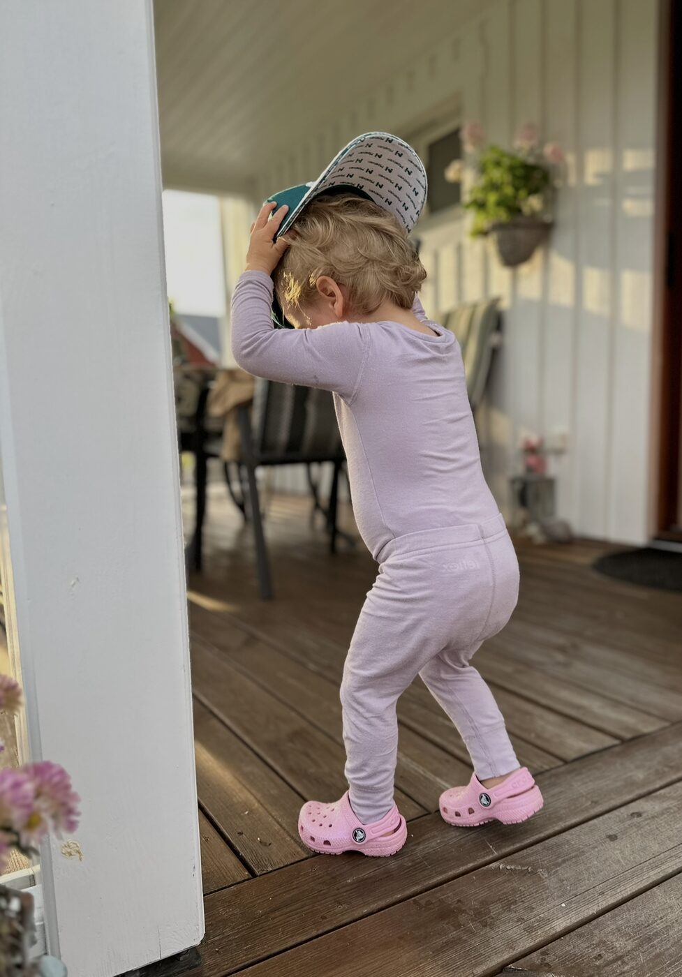 Barn sett bakfra på en veranda i lilla bambusdrakt fra Reflex og rosa Crocs