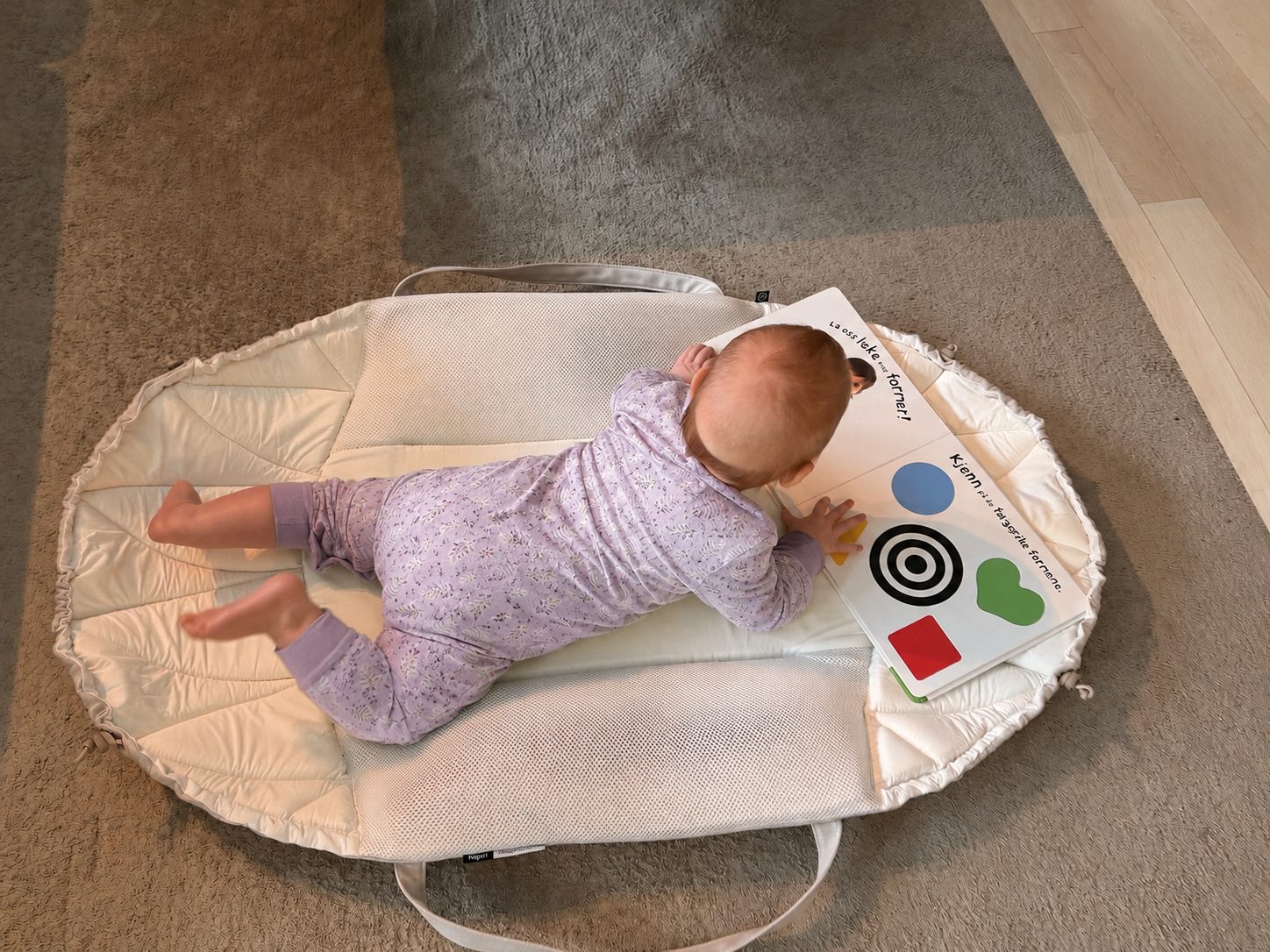 Baby ligger på magen og ser i en bok på Najell SleepCarrier brettet ut som lekematte, sett ovenfra